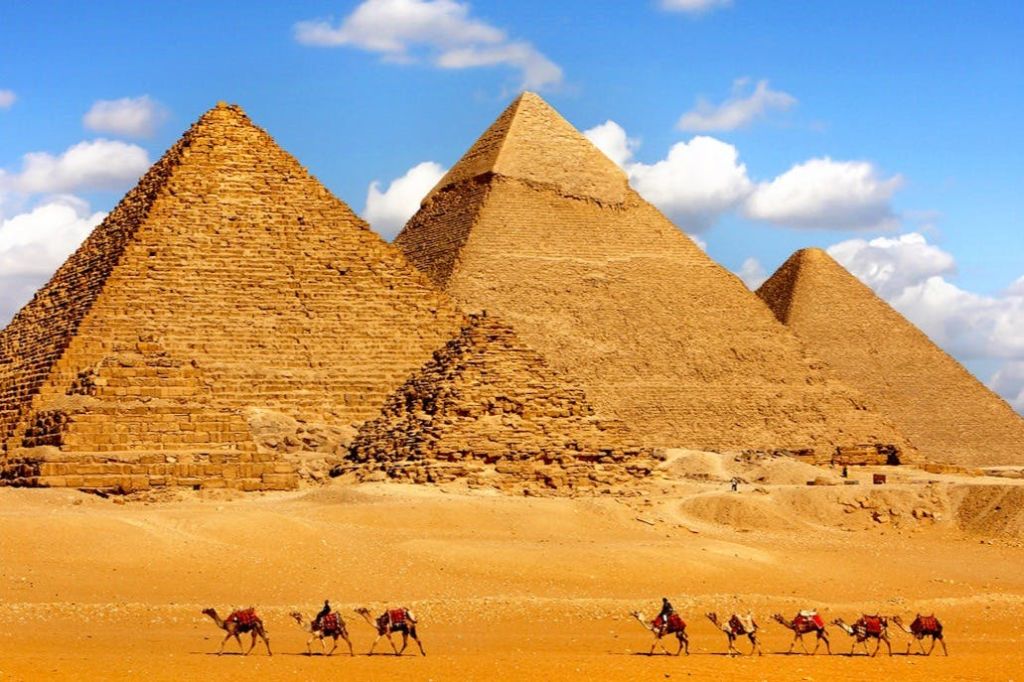 맑고 푸른 하늘 아래 기자의 피라미드가 펼쳐지며 낙타와 기수가 사막을 가로질러 이동합니다.