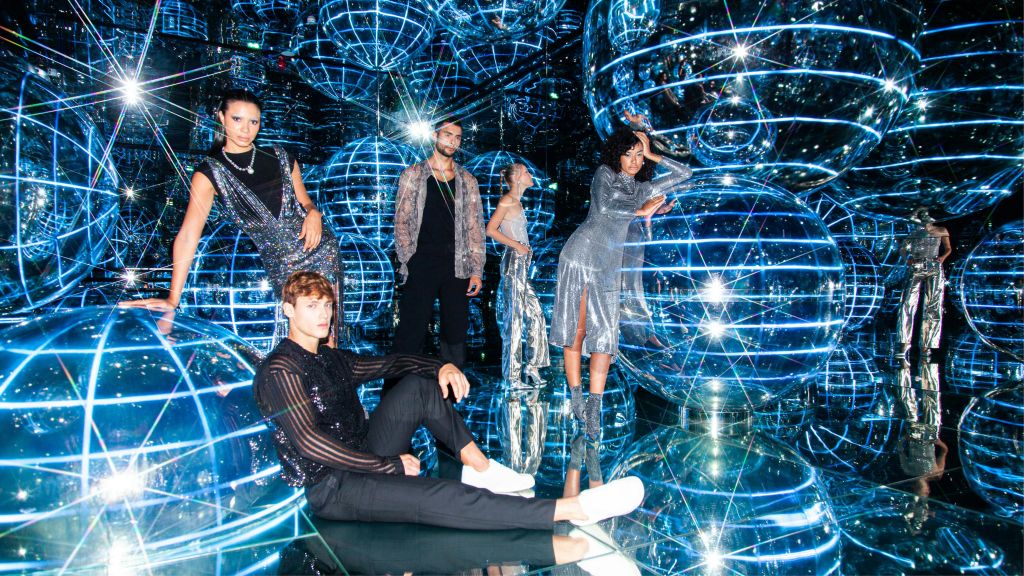 반사되는 표면과 빛나는 파란색 구형 조명을 배경으로 세련되고 반짝이는 의상을 입은 네 사람이 포즈를 취하고 있습니다.