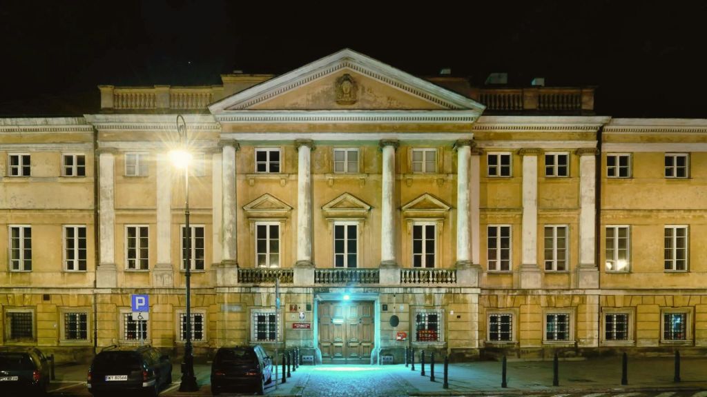 가로등이 켜진 커다란 기둥이 있는 밤의 유서 깊은 건물입니다. 인근에 차량이 주차되어 있습니다.