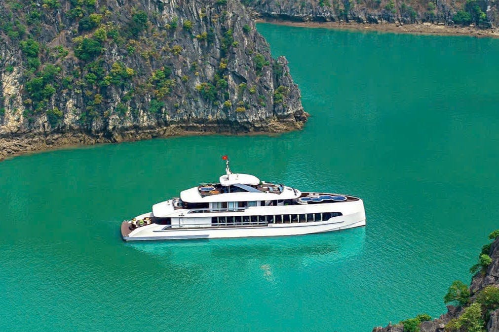 나무로 덮인 바위 절벽 근처의 청록색 물 위에 매끄러운 흰색 요트가 떠 있습니다.