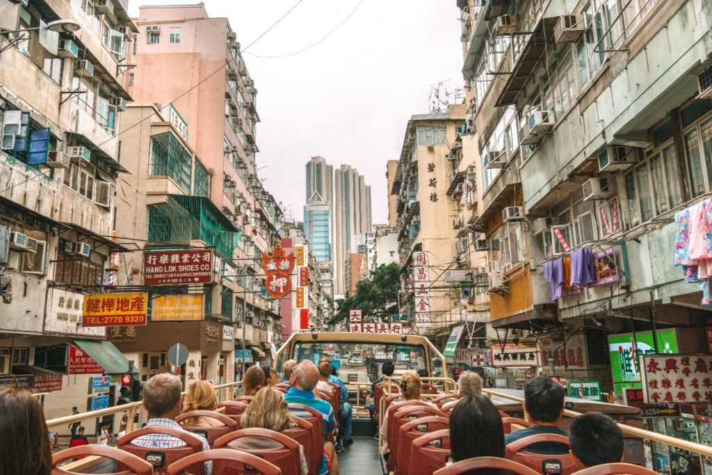 People seated on an open-top bus touring a city street lined with tall urban buildings and storefronts.