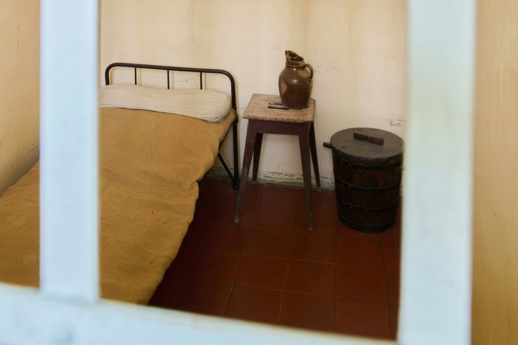 타일 바닥에 금속 프레임 침대, 노란색 담요, 점토 주전자가 있는 나무 의자, 어두운 나무 양동이가 있는 심플한 객실입니다.