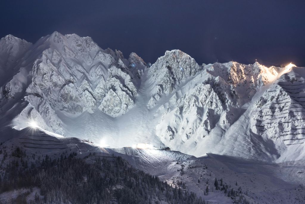 맑은 밤하늘 아래 눈 덮인 산맥, 전경에 밝은 스키장과 소나무가 있습니다.
