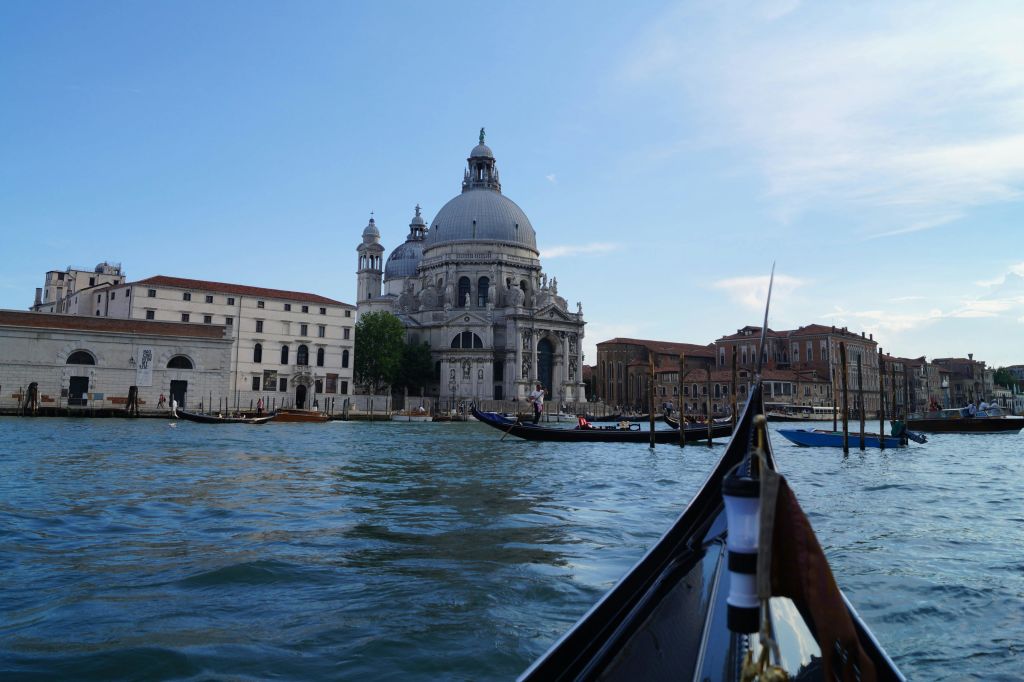 전경에 운하가 있고, 물에 곤돌라가 있고, 머리 위로 맑은 하늘이 보이는 커다란 돔형 건물의 모습.