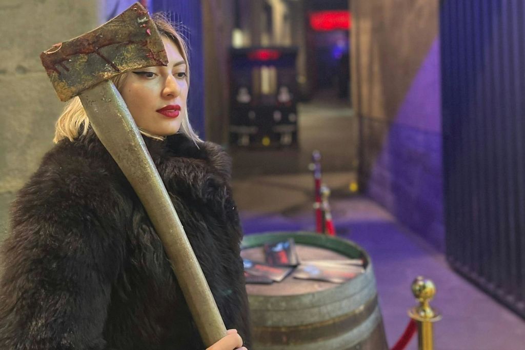 커다랗고 풍화된 도끼를 들고 있는 검은 모피 코트를 입은 긴 금발의 여성. 배경에는 나무 통과 도시 조명이 있습니다.