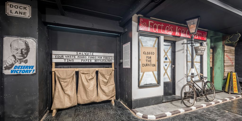 "Closed for the Duration" 표지판이 있는 상점가와 "SALVAGE" 표지판 아래에 통, 종이, 병, 항아리를 보관할 수 있는 재활용 구역이 있습니다.