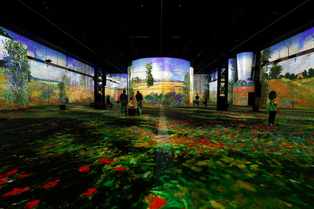 벽과 바닥에 화려하고 인상적인 풍경이 투영된 몰입감 넘치는 예술 작품을 감상하는 사람들.