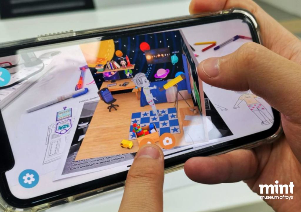 다채로운 물체와 장난감, 체크무늬 러그가 놓인 증강 현실 방을 보여주는 스마트폰을 두 손으로 들고 있습니다.