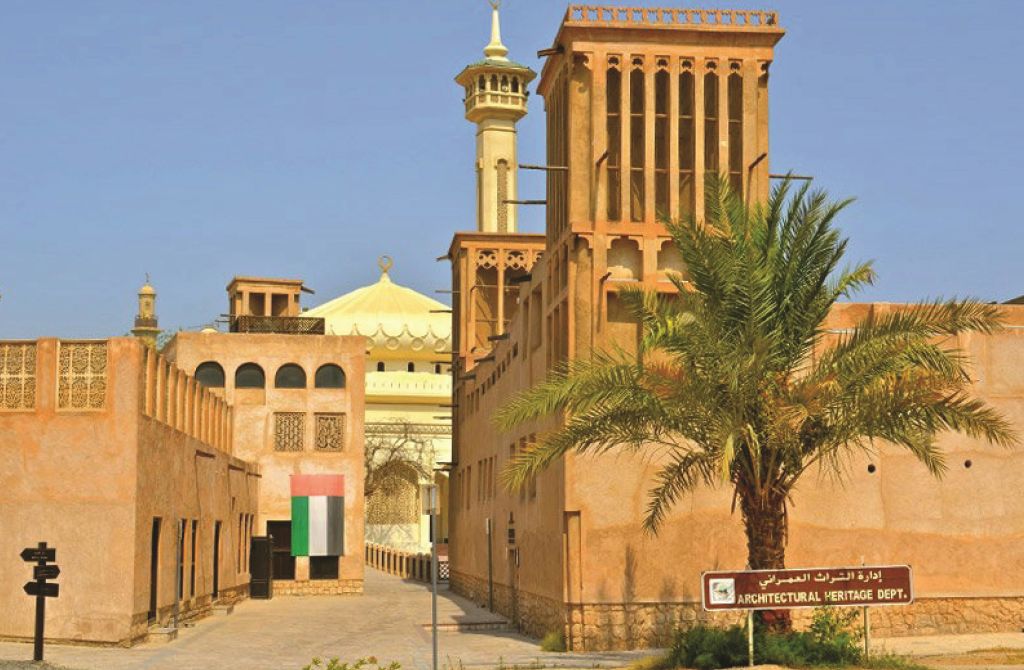 바람의 탑, 야자수, '건축 유산' 표지판이 있는 중동의 전통 건물입니다. 백그라운드에는 모스크가 있습니다.