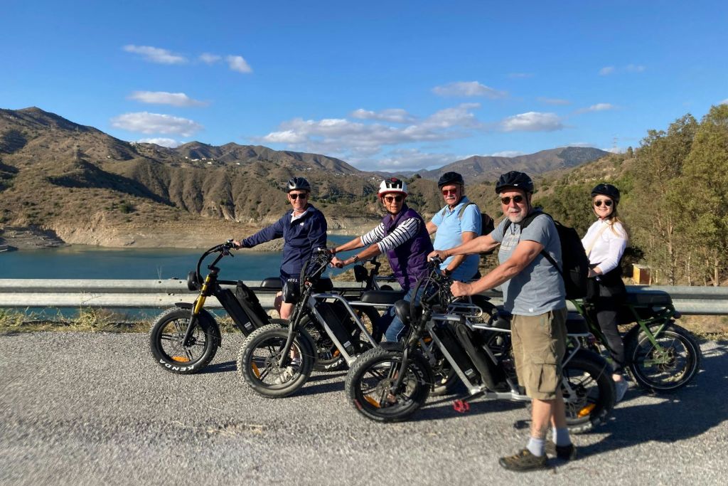 산과 나무, 호수가 배경으로 펼쳐진 아름다운 도로변에는 헬멧을 착용한 5명이 전기 자전거를 타고 서 있습니다.