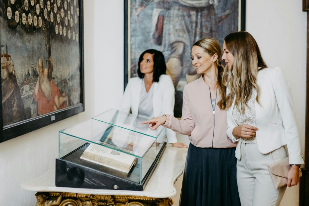 똑똑한 복장을 한 세 명의 여성이 그림 앞에 놓인 유리 케이스에 전시된 책을 감상하고 있습니다.