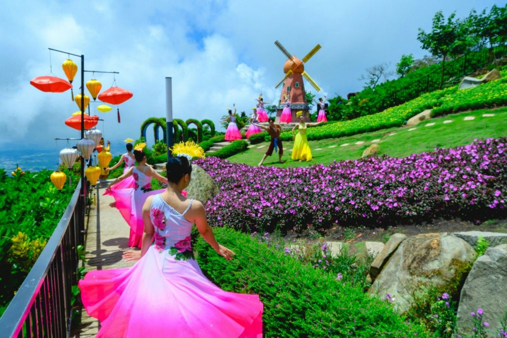 분홍색 드레스를 입은 여성들이 부분적으로 흐린 하늘 아래 꽃이 피고 풍차가 있는 정원 근처에서 춤을 추고 있습니다.