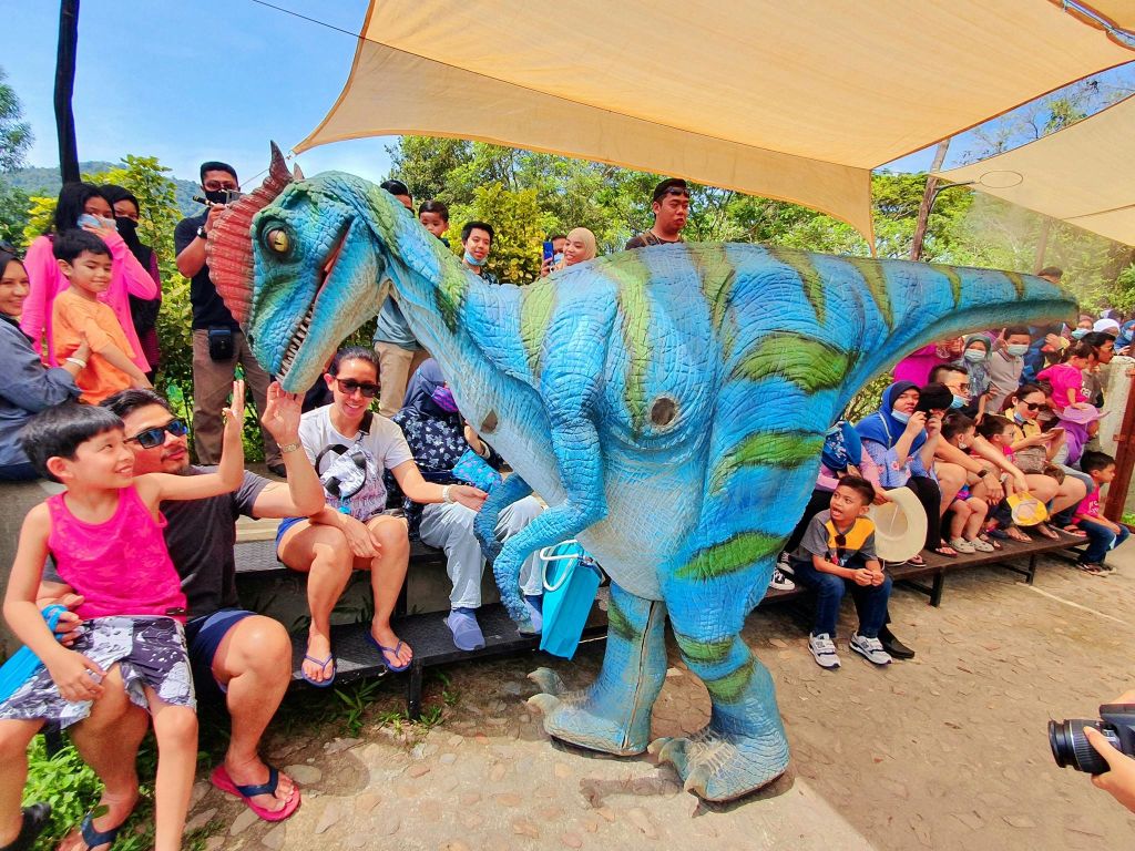 의상을 입은 공룡 엔터테이너가 야외에서 미소 짓는 사람들과 상호 작용하며, 일부 어린이는 그것을 만지기 위해 손을 뻗습니다.