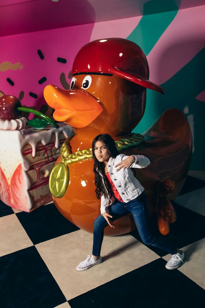 커다란 케이크 소품을 배경으로 모자를 쓴 커다란 오리상 앞에서 장난스럽게 포즈를 취하는 소녀.