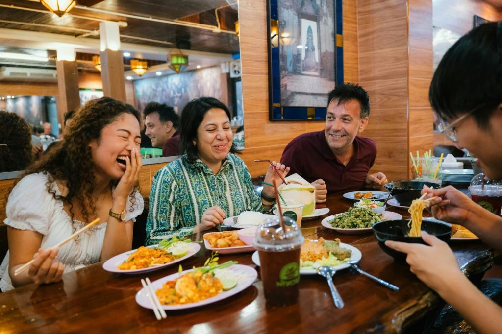 세 사람이 레스토랑의 테이블에 앉아 다양한 요리를 앞에 두고 미소를 짓고 식사를 즐깁니다.