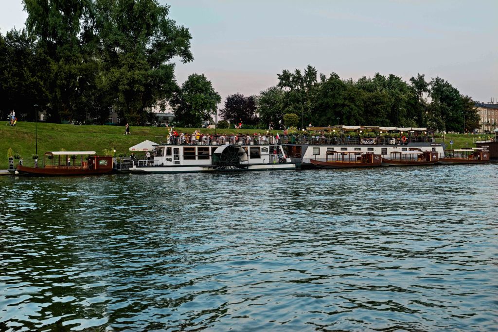 나무가 우거진 강둑을 따라 보트가 정박해 있으며, 사람들이 잔디밭을 거닐고 있습니다. 연한 하늘 아래 잔잔한 물이 흐른다.
