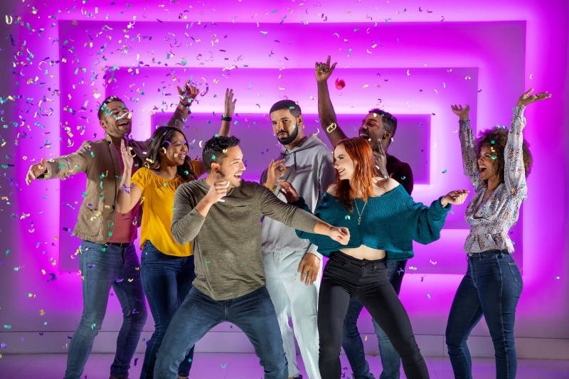 분홍색과 보라색 불빛을 배경으로 6명의 사람들이 콘페티를 들고 파티를 열며 미소 짓고 춤을 추고 있습니다.