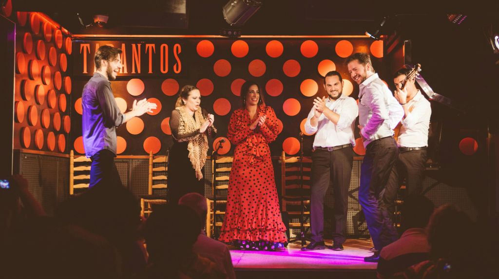 무대에서 주황색 물방울 무늬를 배경으로 공연합니다. 빨간 드레스를 입은 한 댄서가 기타리스트 옆에서 박수를 치고 있습니다.