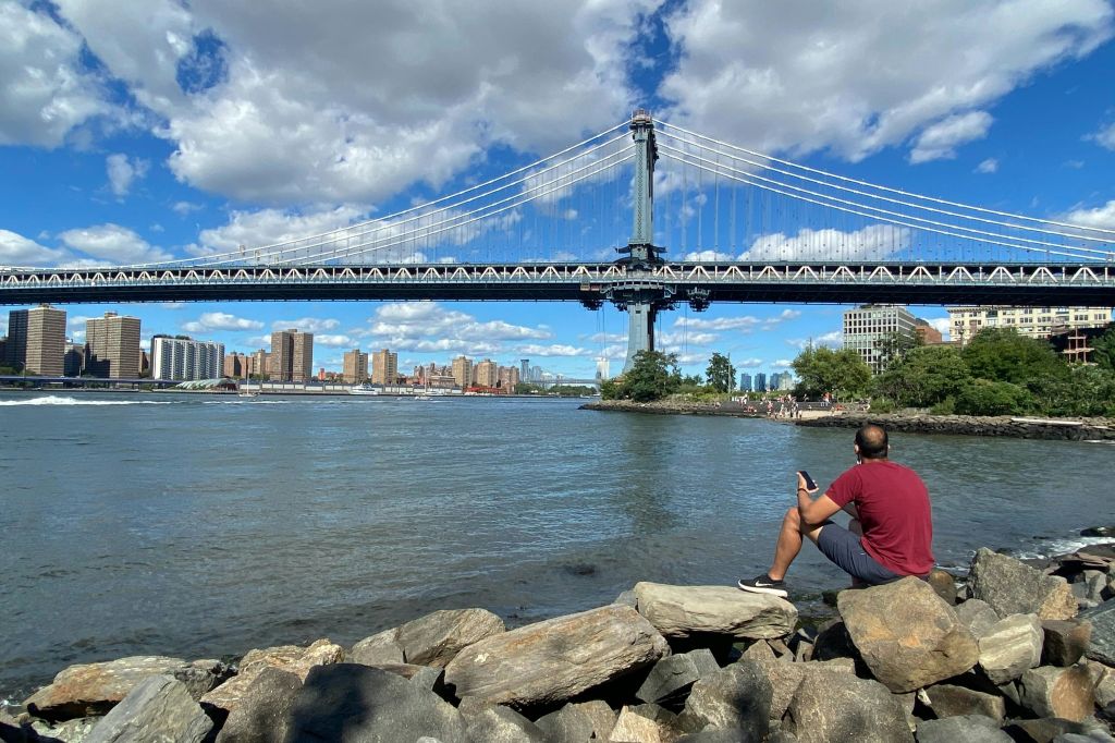 남자는 강가 바위에 앉아 도시의 스카이라인이 있는 현수교와 구름을 배경으로 한 푸른 하늘을 바라보고 있습니다.
