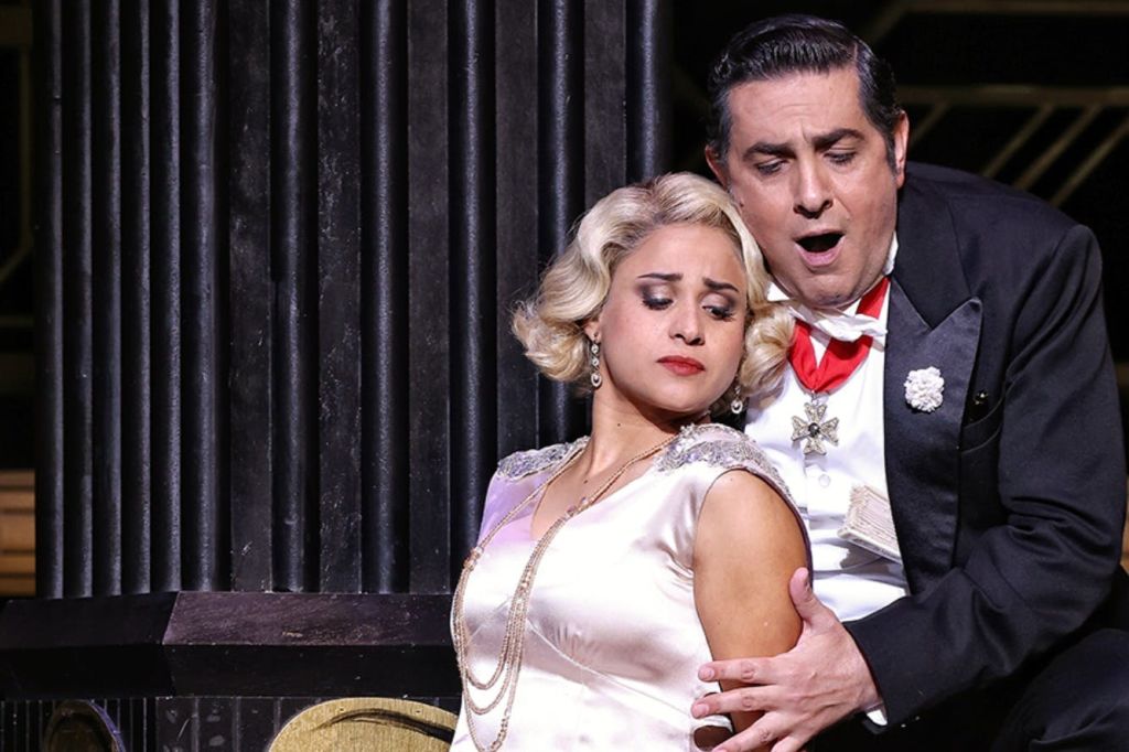 흰색 드레스를 입은 여성과 턱시도를 입은 남성이 무대에서 공연하며 극적인 표정을 짓고 있습니다.