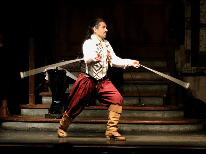 전통 복장을 한 남성이 무대에서 양손으로 리본을 뻗으며 퍼포먼스를 펼치고 있습니다. 계단과 장식용 스탠드가 배경에 있습니다.