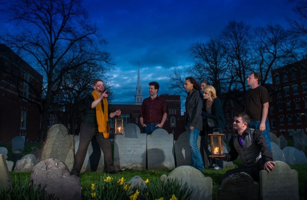 맨발의 나무가 우거진 해질 무렵 묘지의 비석 사이에는 등불을 들고 있는 6명의 무리가 서 있고, 그 뒤로 교회의 첨탑이 있습니다.