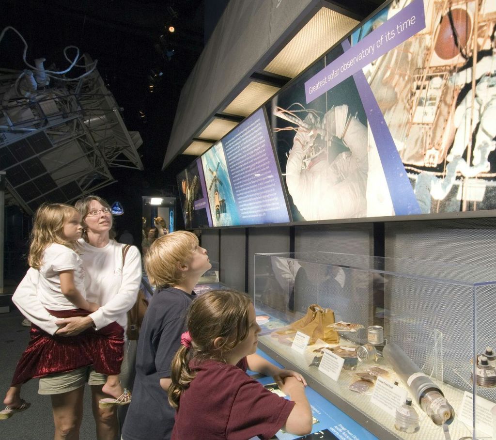 한 여성과 세 명의 어린이가 박물관에서 우주 전시물과 유물을 관찰하고 있으며, 인공위성 모형이 배경으로 전시되어 있습니다.