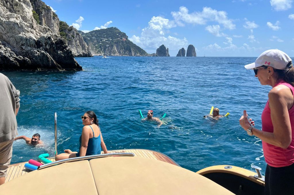 두 사람이 수영장 국수를 들고 바다에 떠 있으며, 보트에는 뚜껑과 탱크탑을 찬 여성이 서 있습니다. 바위 절벽과 바다 더미가 배경입니다.