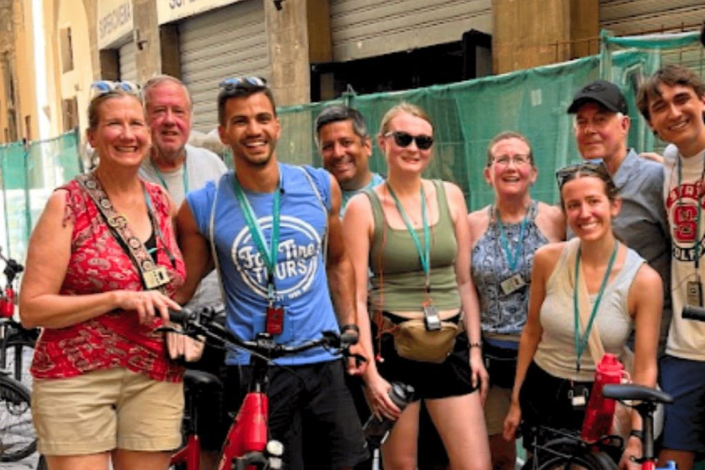 한 무리의 사람들이 도시 거리에서 자전거를 타고 미소를 짓고 포즈를 취하고 있습니다. 건물 정면이 한눈에 들어옵니다.