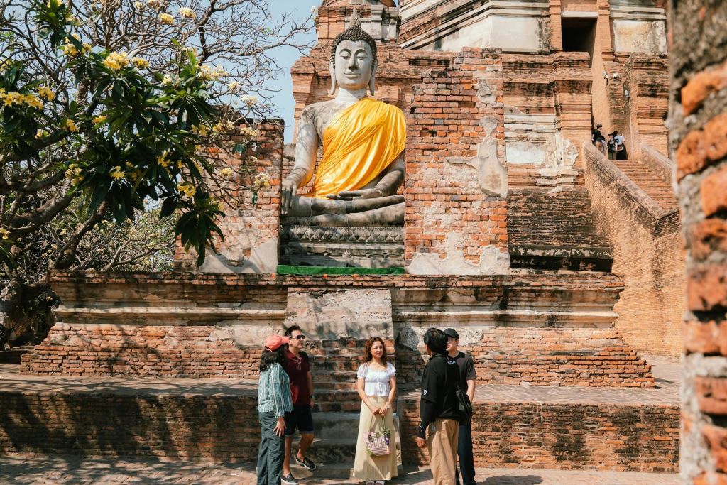 한 무리의 사람들이 오래된 벽돌 구조물과 나무를 배경으로 노란색으로 장식된 커다란 불상 근처에 서 있습니다.