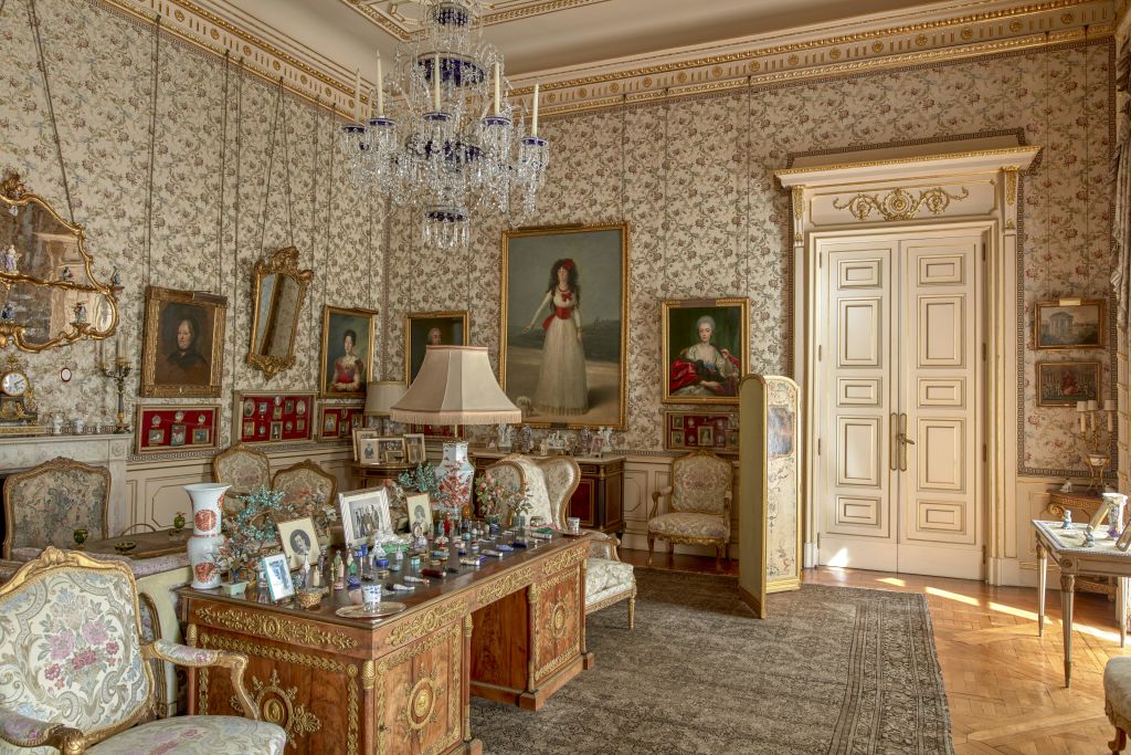 꽃무늬 벽지, 커다란 샹들리에, 벽에 걸린 그림, 다양한 장식품이 놓인 나무 책상이 있는 화려한 객실.