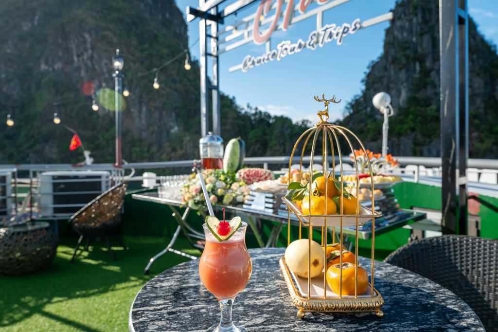 열대 음료, 황금 케이지의 과일, 뷔페 테이블, 줄 조명, 산의 풍경을 배경으로 한 야외 식사 공간.