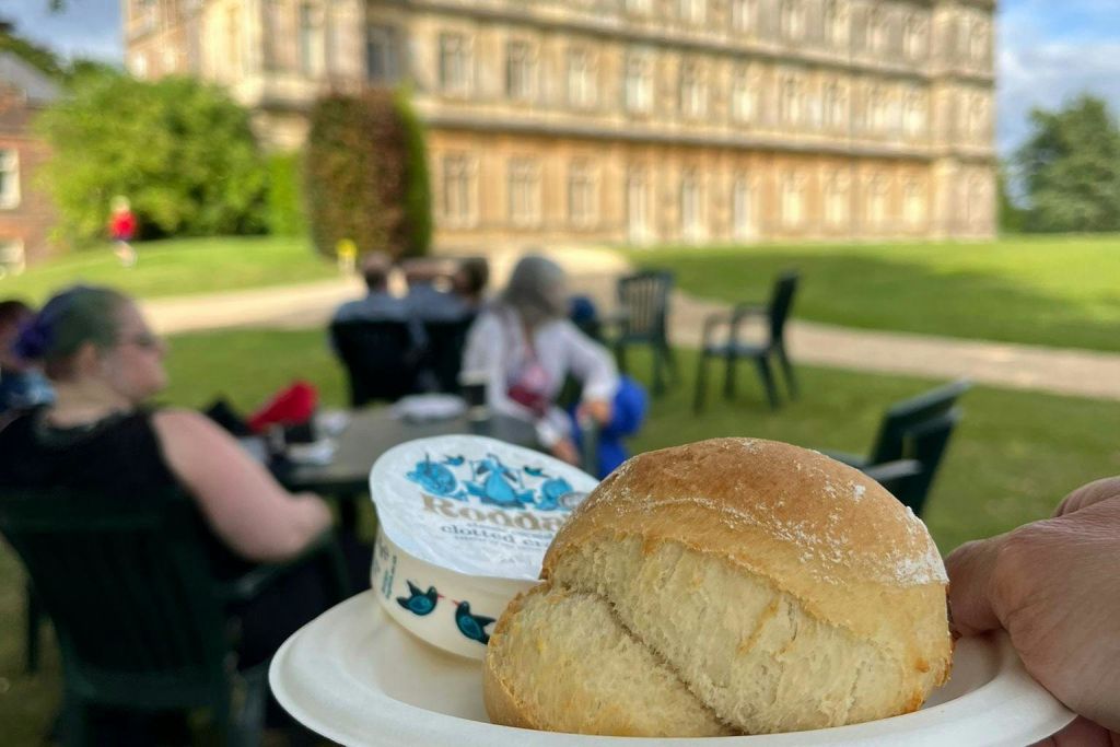 빵 롤과 스프레드 용기가 담긴 접시가 전면에 놓여 있으며, 큰 건물 근처에 사람들이 야외에 앉아 있습니다.