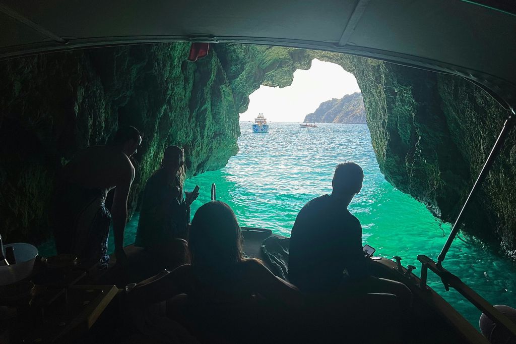 청록색 물과 보트가 보이는 동굴 안에 앉아 있는 사람들, 햇살이 동굴 입구를 비춥니다.