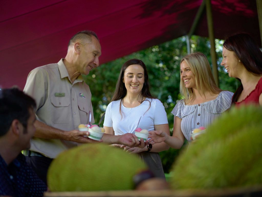 세 사람이 와플 콘에서 아이스크림을 들고 웃고 있으며, 커다란 녹색 과일 근처의 마룬 캐노피 아래 야외에 서 있습니다.