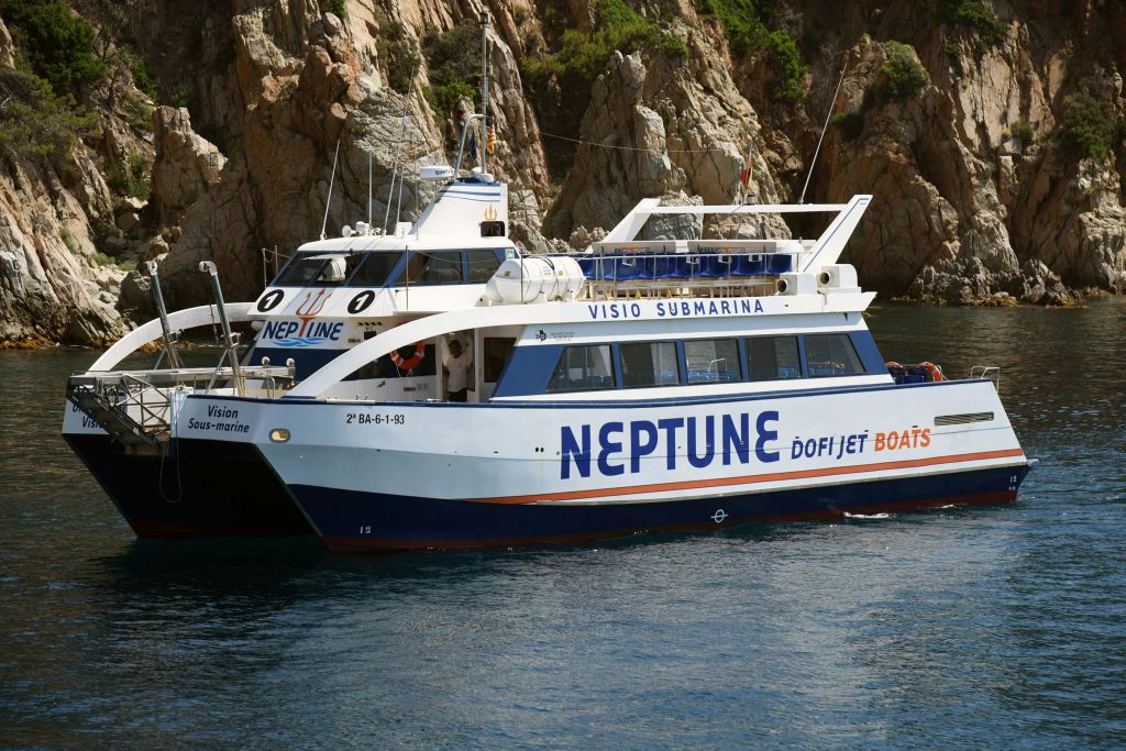 물가의 바위 절벽 근처에 "NEPTUNE" 과 "VISIO SUBMARINA" 라고 표시된 흰색과 파란색 보트가 있습니다.