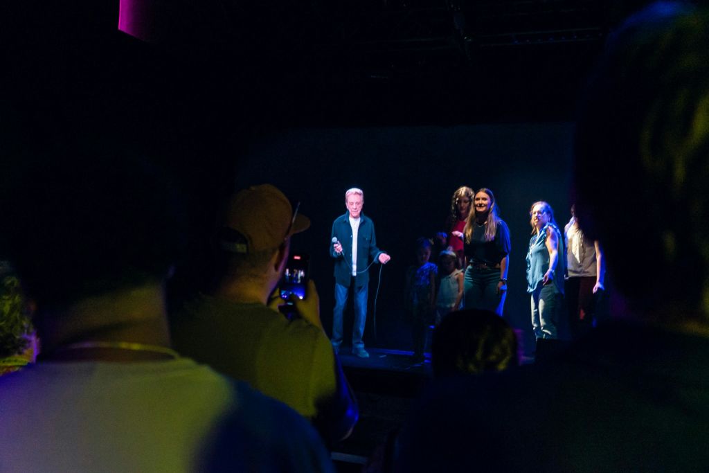 한 무리의 사람들이 어둡게 빛나는 무대에 서고, 다른 사람들은 지켜보며 사진을 찍습니다.