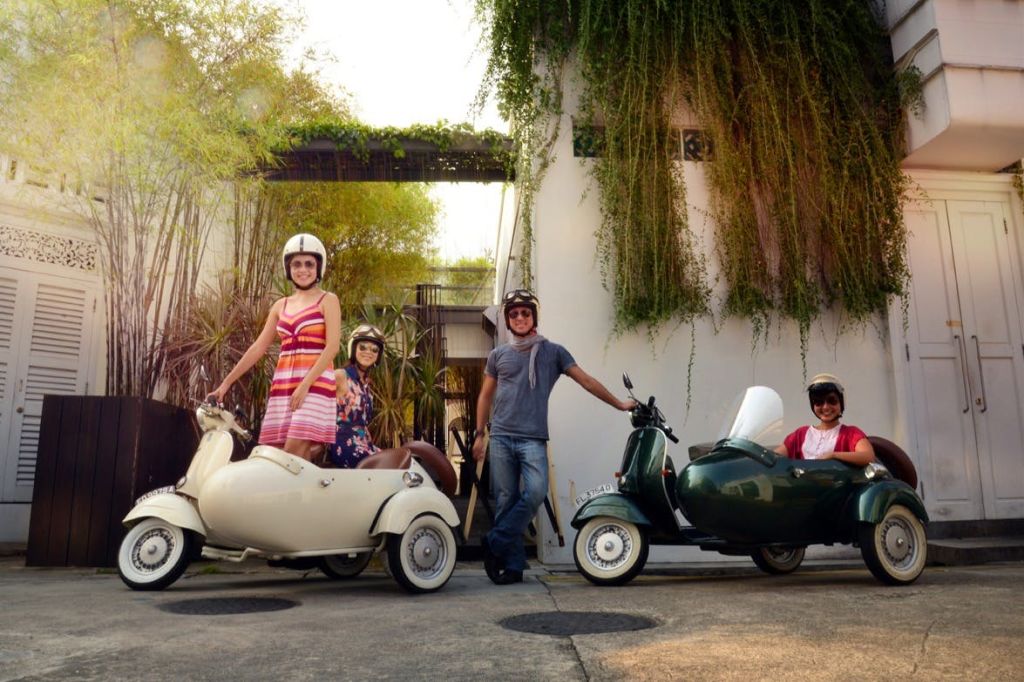헬멧을 쓴 세 사람이 빈티지 전동 스쿠터 옆에 서 있습니다. 전동 스쿠터 한 대에 사이드카가 있습니다. 배경에는 식물과 흰색 건물이 있습니다.