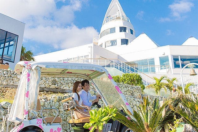 분홍색 하트로 장식된 골프 카트에 하얀 건물과 푸른 하늘을 배경으로 커플이 앉아 있습니다.