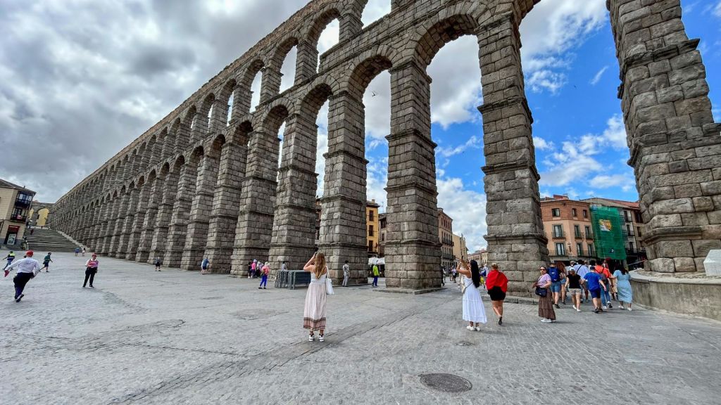 도시 광장의 부분적으로 흐린 하늘 아래, 아치가 있는 고대 석조 수로 근처를 걷는 사람들.