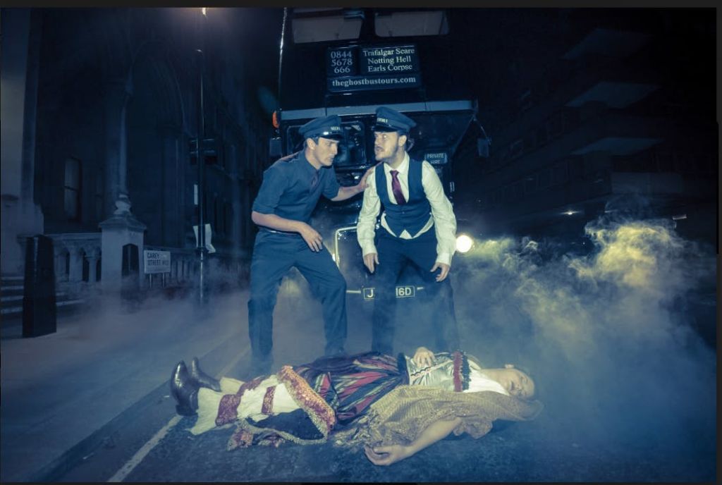 제복을 입은 두 남자가 버스 옆에 서서 밤에 안개가 자욱한 거리에서 바닥에 누워 있는 역사적인 옷을 입은 사람을 바라보고 있습니다.