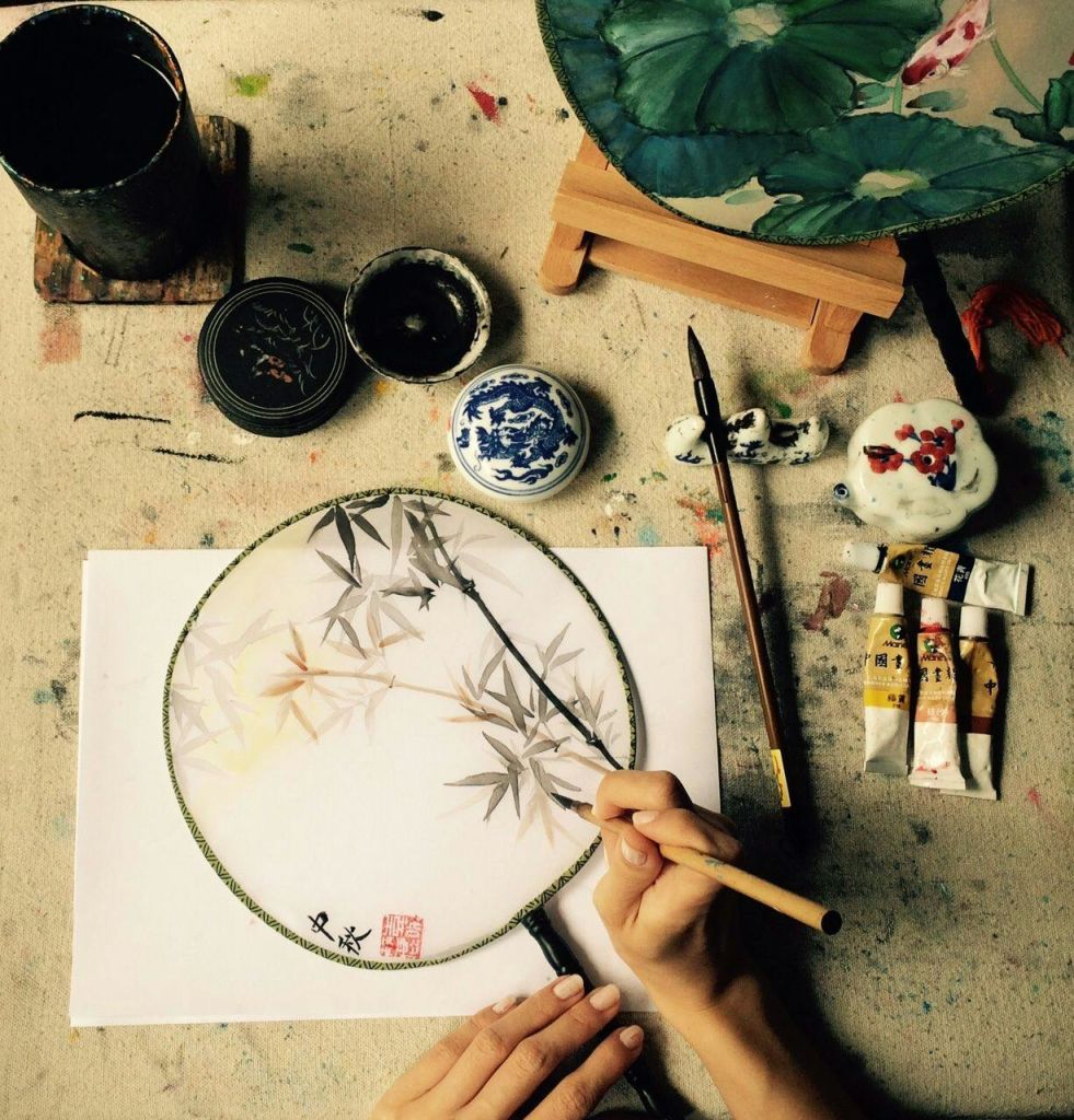 원형 선풍기에 대나무를 직접 그려보세요. 근처에는 페인트 튜브, 브러시, 잉크 냄비, 꽃무늬 디자인의 완성된 선풍기가 있습니다.