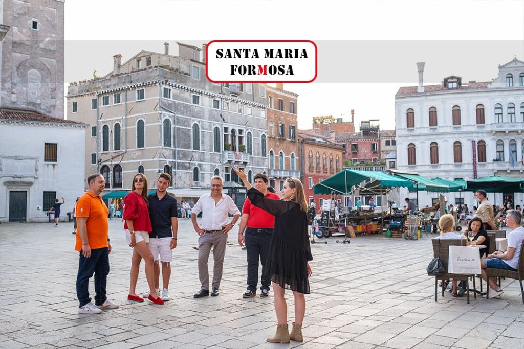 검은 드레스를 입은 가이드가 오래된 건물 앞의 유서 깊은 광장에 서 있는 5명의 관광객에게 무언가를 설명하고 있습니다.