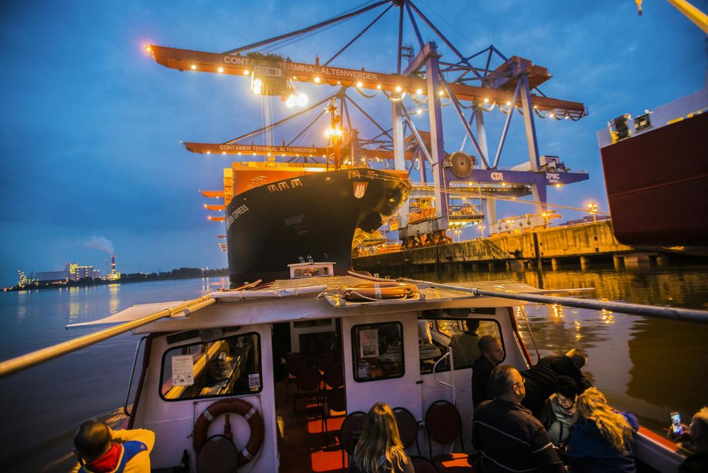 해질 무렵 대형 화물선 근처에서 보트를 타는 사람들과 물가의 컨테이너 터미널에서 크레인을 타는 사람들.