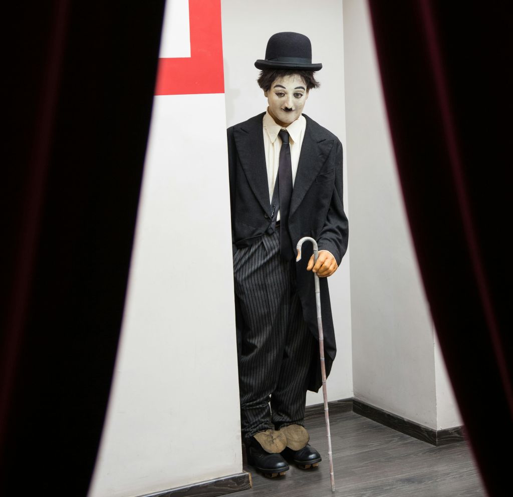 벽 뒤에 부분적으로 숨겨져 있는 찰리 채플린의 옷을 입은 사람이 빨간색 커튼 사이로 보이는 지팡이를 들고 있습니다.