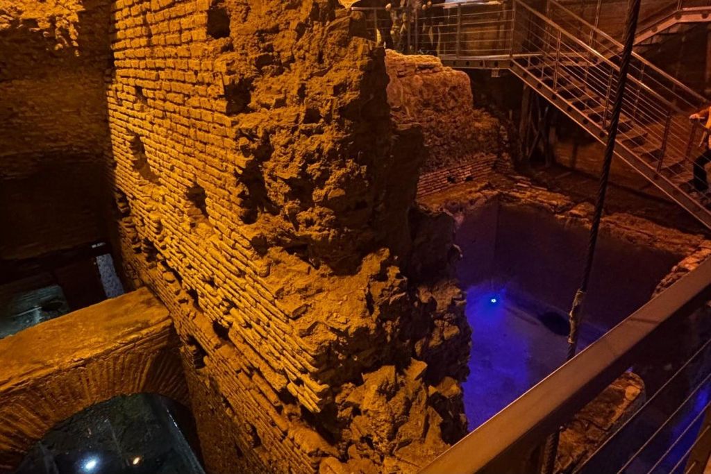 고대 벽돌 벽에는 따뜻한 조명이 켜져 있으며, 아래쪽에는 파란색 조명이 켜져 있고 배경에는 현대적인 난간이 있습니다.