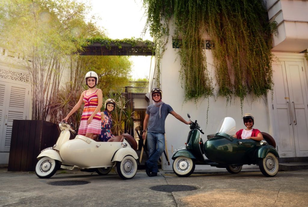 벽에 녹음이 우거진 건물 앞에 모터 스쿠터를 든 세 사람이 서 있습니다. 한 사람은 사이드카를 타고 서 있습니다.