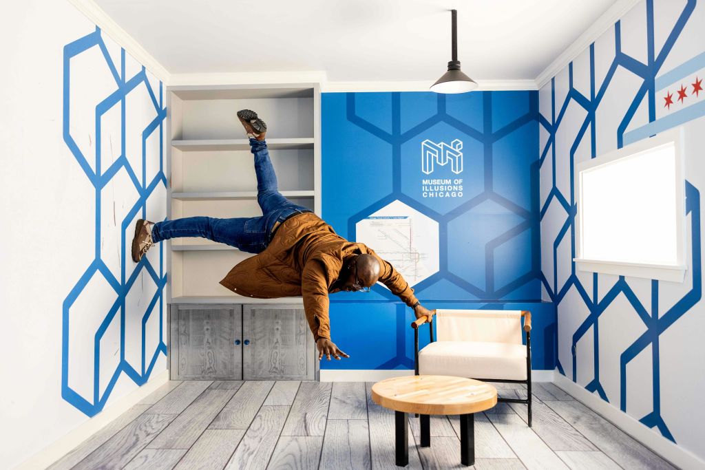 한 사람이 파란색과 흰색의 기하학적 패턴과 '시카고 환상 박물관' 표지판이 있는 방에 수평으로 떠 있는 것으로 보입니다.