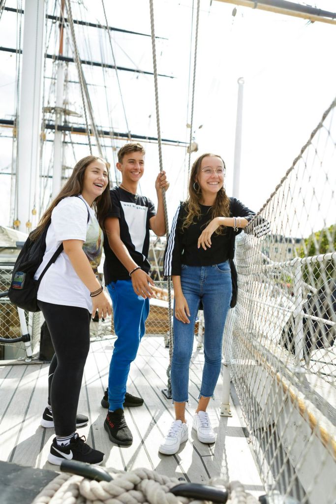 도킹된 배에 서서 밧줄을 잡고 리깅으로 둘러싸인 미소 짓는 청소년 세 명.