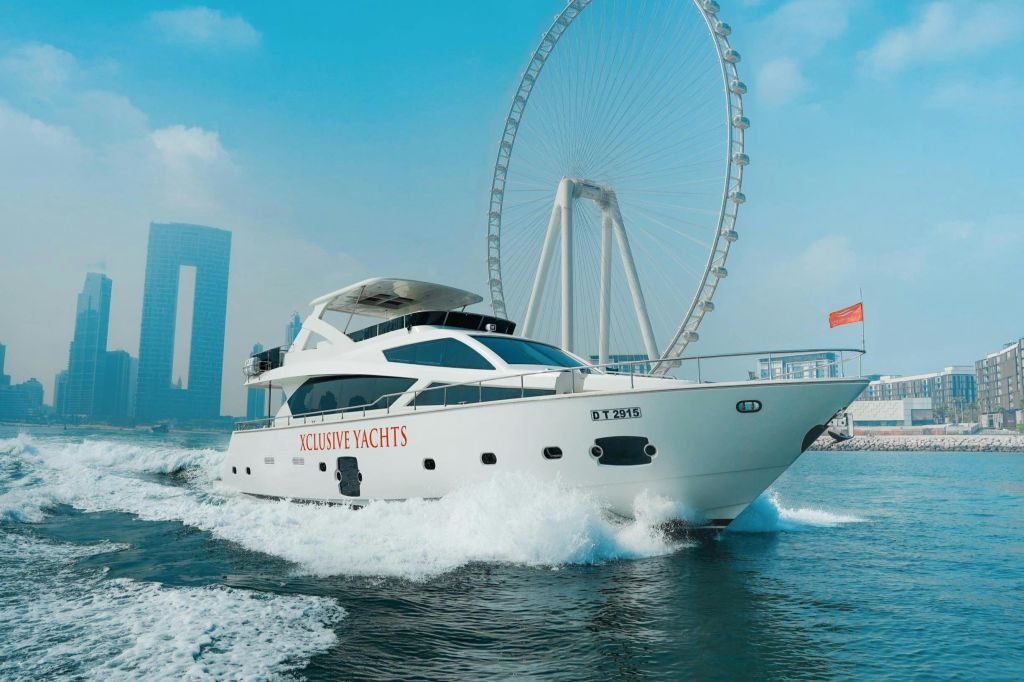 대형 대관람차와 고층 빌딩을 배경으로 물 위를 항해하는 사이드 돛에 "익스클루시브 요트 (Xclusive Yachts)" 라는 이름의 흰색 요트가 있습니다.